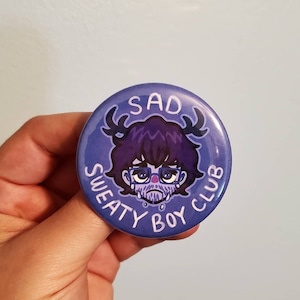 May include: A purple button with a cartoon illustration of a sad character with antlers, glasses, and a beard. The text "SAD SWEATY BOY CLUB" surrounds the character.