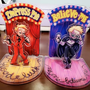 May include: Two acrylic character stands featuring cartoon characters with red and blue backgrounds. The first stand features a red devil character with the text "Impress Me" above. The second stand features a blue character with the text "Believe Me" above.