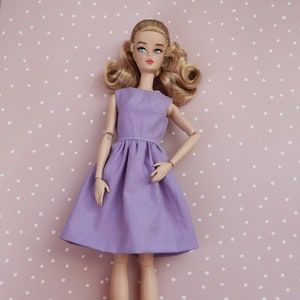 Könnte beinhalten: Eine Puppe mit blonden Haaren und blauen Augen, die ein lilafarbenes, ärmelloses Kleid mit einem gerafften Rock trägt.