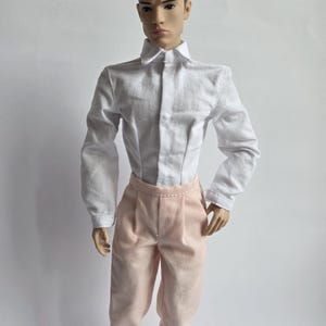 May include: A fashion doll with dark brown hair styled in a side part, wearing a long-sleeved white button-down shirt with a pointed collar and light pink trousers featuring a pleated waistline and a visible center seam down the front of the legs.