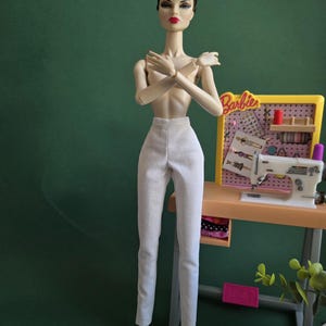 May include: A fashion doll with brown hair styled in an updo, wearing plain white trousers. The doll stands next to a miniature sewing station featuring a white sewing machine, a yellow pegboard with the text Barbie, and various craft accessories.
