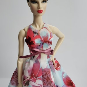 May include: A fashion doll wearing a pink, blue and white floral print dress with a burgundy satin ribbon tied around the waist.