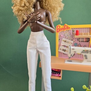May include: A doll with dark skin and voluminous blonde curly hair wearing high-waisted white trousers. The doll stands next to a miniature sewing station featuring a white sewing machine, a pink pegboard with the Barbie logo, and various accessories.