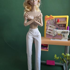 May include: A blonde fashion doll with long wavy hair wearing white trousers, standing in front of a miniature sewing station. The station features a yellow pegboard with the word Barbie, a white sewing machine, and various small craft supplies.