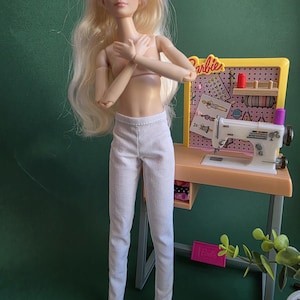 May include: A blonde doll with long hair wearing plain white trousers, standing in front of a miniature yellow sewing desk. The desk features a white sewing machine, a pink pegboard with various craft accessories, and the Barbie logo in yellow.