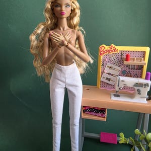 May include: A fashion doll with long wavy blonde hair and tan skin, wearing white trousers. Beside the doll is a miniature sewing station featuring a white sewing machine, a yellow pegboard with the Barbie logo, and a small shelf with thread.