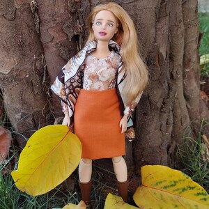 May include: A doll wearing an orange skirt, a patterned top, and a scarf with brown and white designs. The doll has long blonde hair and brown boots. The doll is holding a large yellow leaf.