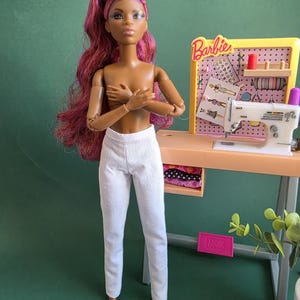 Op de afbeelding: Een pop met lang golvend magentakleurig haar en een bruine huid draagt een witte broek. Naast de pop staat een miniatuur naaiwerkstation met een witte naaimachine, een geel prikbord met de tekst Barbie en diverse kleine naai-accessoires.