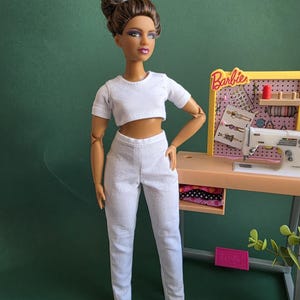 Könnte beinhalten: Eine Puppe mit braunem Haar in einem hohen Dutt, bekleidet mit einem weißen kurzärmeligen bauchfreien Oberteil und einer passenden weißen langen Hose. Daneben steht eine Miniatur-Nähstation mit einer weißen Nähmaschine und dem Text Barbie.