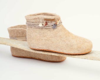 May include: A pair of beige felted wool slippers. The ankle-high slippers have a decorative band with small floral accents. The soles have an orange trim. The slippers are displayed on a piece of light-colored wood.