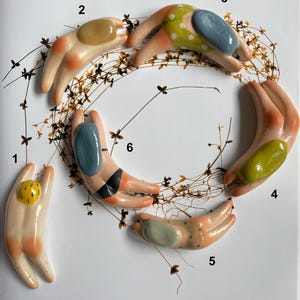 Puede incluir: Una colección de seis esculturas de manos de cerámica, cada una con un diseño y una paleta de colores únicos. Las manos están dispuestas en un patrón circular, con detalles que incluyen acentos pintados y superficies texturizadas. La obra de arte está sobre un fondo blanco con delicados tallos de plantas.