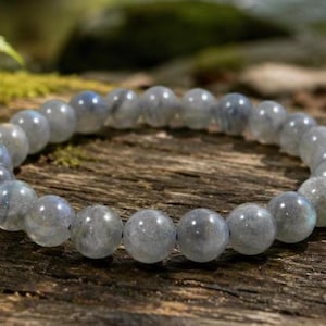 May include: A beaded bracelet made of round, gray labradorite stones. The bracelet is displayed on a weathered wooden surface with hints of green moss and foliage in the background. The stones have a subtle iridescence.