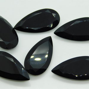 May include: Six black pear-shaped gemstones with a faceted cut. The gemstones are arranged in a semi-circle on a white surface.