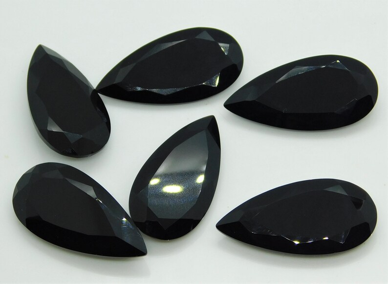 May include: Six black pear-shaped gemstones with a faceted cut. The gemstones are arranged in a semi-circle on a white surface.