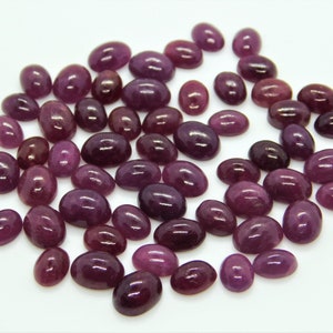 Könnte beinhalten: Eine Sammlung oval geschliffener Edelsteine in verschiedenen tiefroten und violetten Farbtönen. Die polierten Steine sind auf einer weißen Oberfläche verstreut und zeigen ihre glatten, reflektierenden Oberflächen. Die Steine variieren in Größe und Farbe.