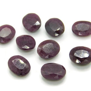 Può includere: Dieci pietre preziose di rubino di forma ovale e sfaccettate in varie tonalità di viola e rosso. Le pietre preziose sono disposte in uno schema sparso su uno sfondo bianco.