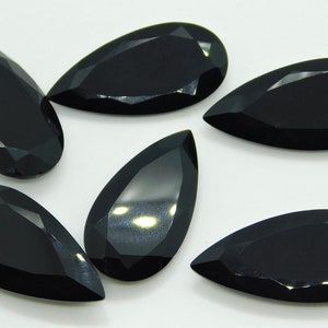 May include: Six black pear-shaped gemstones with a faceted cut. The gemstones are arranged in a semi-circle on a white surface.