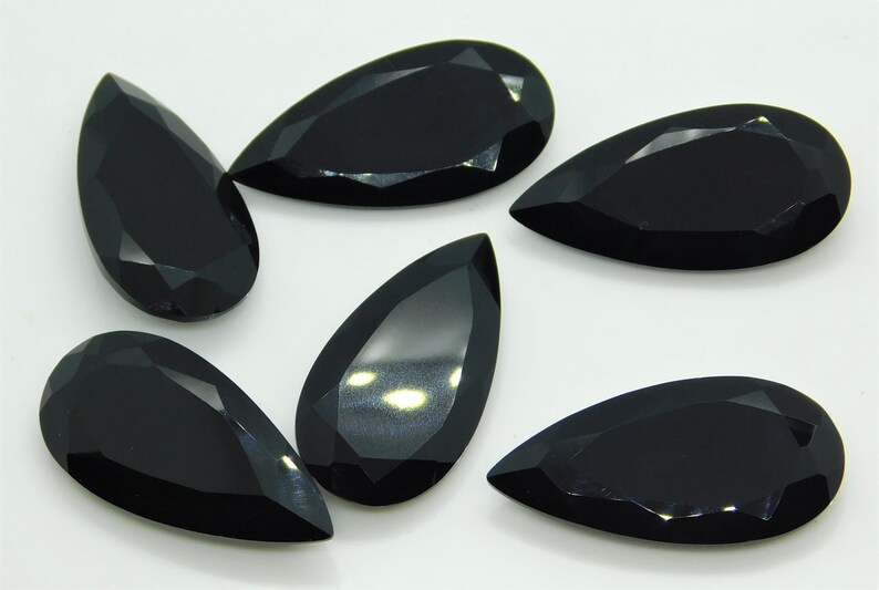 May include: Six black pear-shaped gemstones with a faceted cut. The gemstones are arranged in a semi-circle on a white surface.