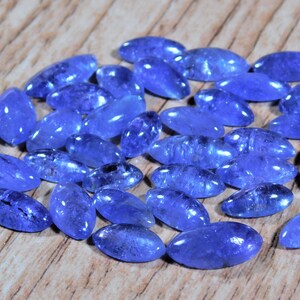 May include: A collection of oval-shaped, faceted, blue gemstones. The gemstones are arranged in a loose pile on a wooden surface.