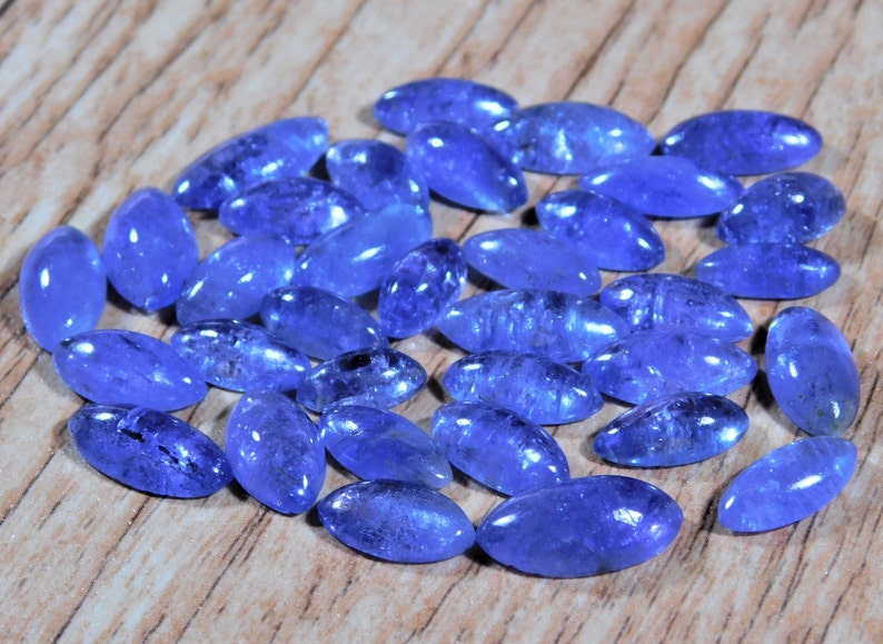 May include: A collection of oval-shaped, faceted, blue gemstones. The gemstones are arranged in a loose pile on a wooden surface.