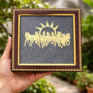 Può includere: Un'opera d'arte incorniciata con una raffigurazione dorata di cavalli che corrono sotto un sole. L'opera è ambientata su uno sfondo scuro e strutturato e incorniciata in legno marrone scuro con accenti dorati. La cornice è quadrata.