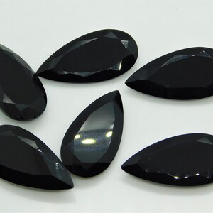 May include: Six black pear-shaped gemstones with faceted edges. The gemstones are arranged in a semi-circle on a white surface.