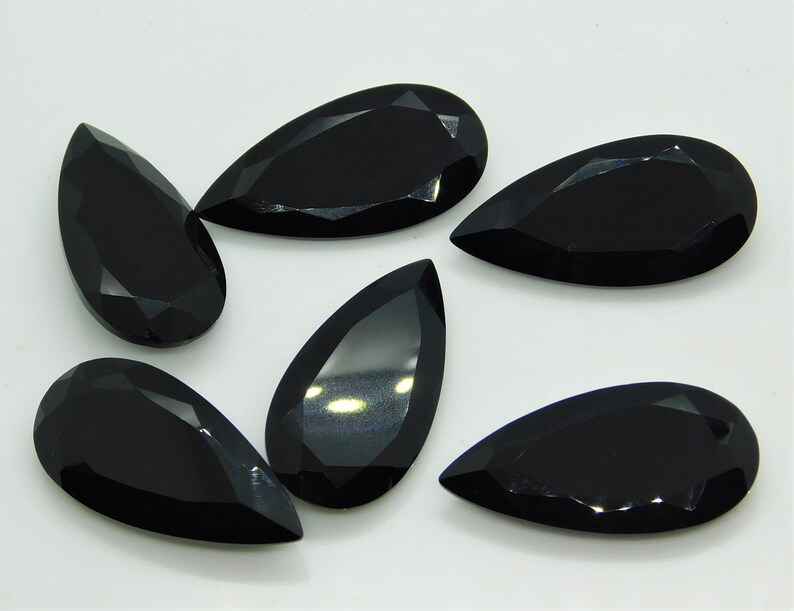 May include: Six black pear-shaped gemstones with faceted edges. The gemstones are arranged in a semi-circle on a white surface.