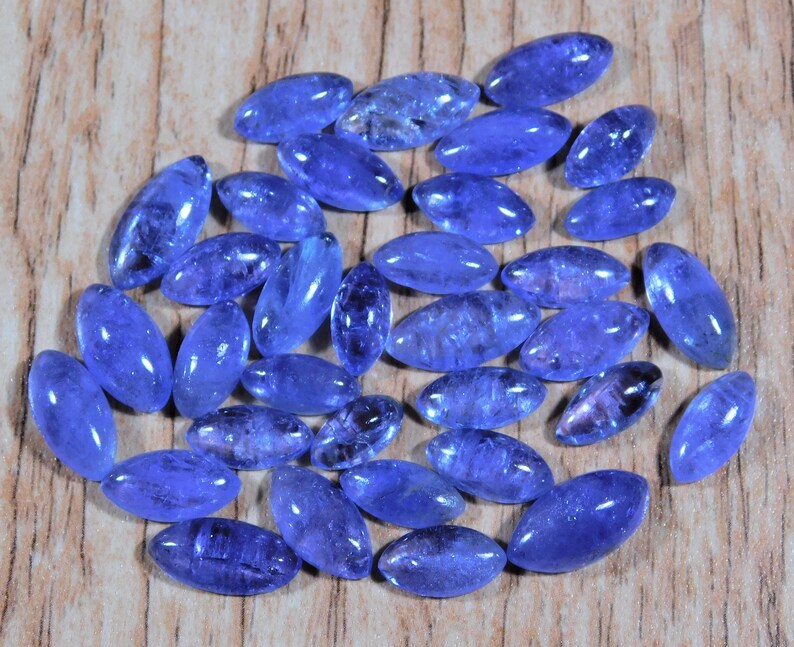 May include: A collection of oval-shaped, faceted, blue gemstones. The stones are arranged in a loose pile on a wooden surface.