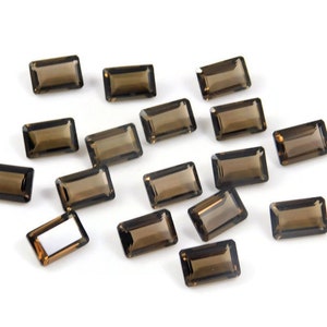 Puede incluir: Una colección de gemas de cuarzo ahumado rectangulares. Cada gema tiene un rico tono marrón y un corte rectangular. Las piedras están dispersas sobre una superficie blanca.