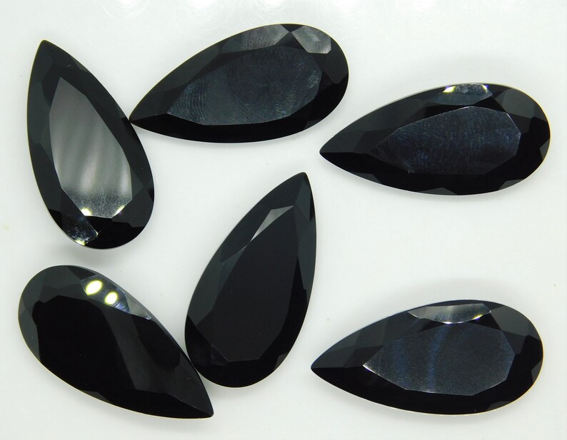 May include: Six black pear-shaped gemstones on a white background. The gemstones are faceted and have a glossy finish.