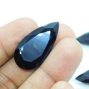 May include: A black pear-shaped gemstone with a faceted cut. The gemstone is held in a hand, and the background is white.