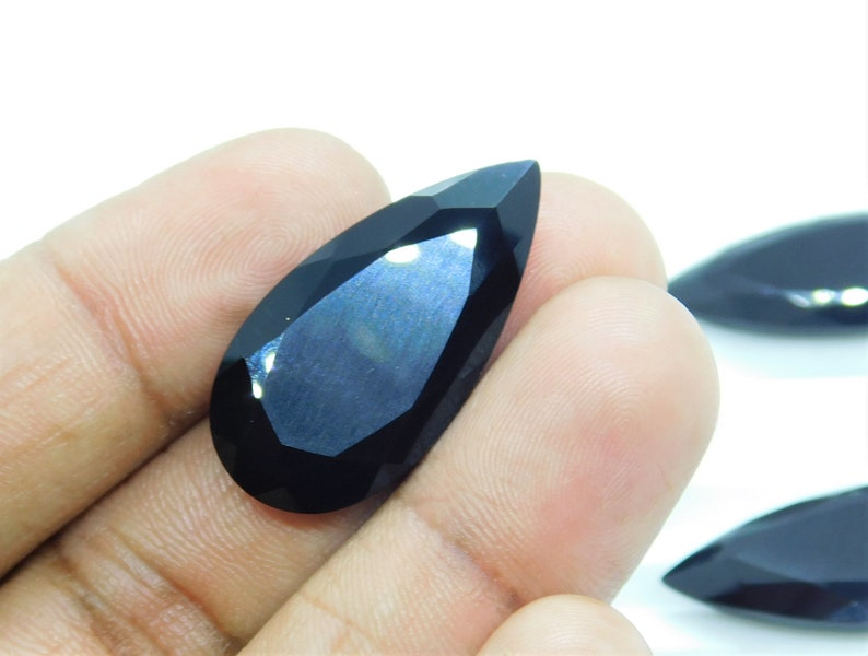 May include: A black pear-shaped gemstone with a faceted cut. The gemstone is held in a hand, and the background is white.