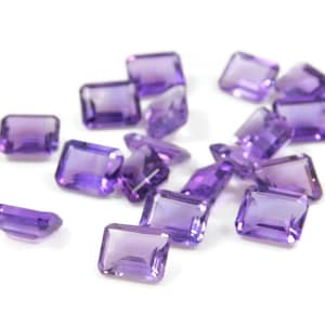 Puede incluir: Un conjunto de gemas de amatista rectangulares y facetadas. Las gemas son de color púrpura intenso y están dispersas sobre una superficie blanca. Las facetas capturan la luz, creando reflejos y sombras. Las gemas varían ligeramente en tamaño y forma.