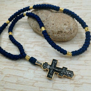 Può includere: Una corda di preghiera annodata blu navy con accenti color oro e un ciondolo a croce dettagliato. La croce presenta un design nero e argento con figure religiose. La corda è drappeggiata su una pietra.