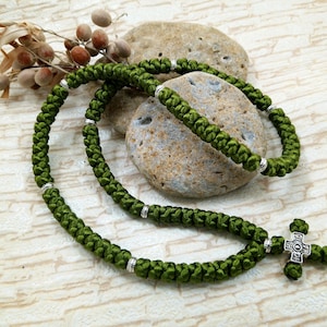May include: Olive green knotted prayer rope with a silver cross pendant and silver accents. The rope is draped over two stones, with a sprig of dried berries in the background. A religious accessory.