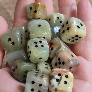 May include: A handful of gemstone dice in shades of green, beige, and brown. Each die features black dots representing numbers. The dice are held in a hand, highlighting their unique patterns and textures. The dice are approximately 1.5 cm.