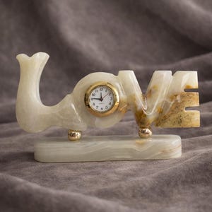 Puede incluir: Un reloj decorativo de mármol con la forma de la palabra "LOVE". La esfera del reloj es dorada con números negros. El mármol es blanco con vetas marrones y beige, y el reloj descansa sobre una base rectangular.