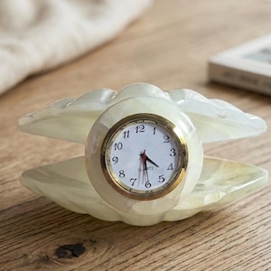 May include: A quartz clock in a shell design. The clock face is white with black numerals and gold trim. The shell is a creamy, translucent colour, resting on a wooden surface.