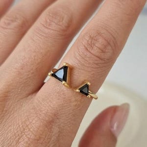 Puede incluir: Un anillo dorado con dos gemas triangulares negras. El anillo tiene un diseño de banda abierta, con un triángulo en la parte superior y otro más pequeño en el lateral. Las gemas están engastadas en un metal dorado.