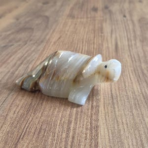 Puede incluir: Una taza de cerámica blanca con asa negra y el texto "But First, Coffee" en negrita negra. La taza se encuentra sobre un fondo borroso, sugiriendo un ambiente acogedor.