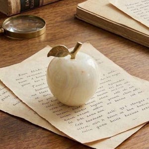 Op de afbeelding: Een decoratieve witte marmeren appel met een gouden steel en blad, geplaatst op oud papier met handschrift. Een vergrootglas en boeken zijn zichtbaar, wat een vintage esthetiek creëert. Decoratief item.