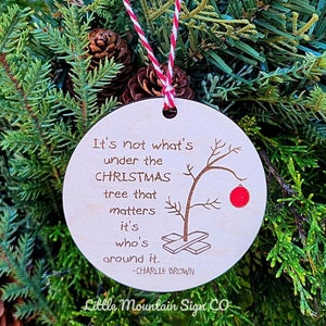 May include: A round wooden Christmas ornament with a quote from Charlie Brown from the Peanuts cartoon. The quote reads "It's not what's under the Christmas tree that matters it's who's around it." The ornament features a small illustration of a Charlie Brown Christmas tree.