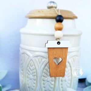 May include: A white ceramic jar with a wooden lid and a brown wooden coffee cup charm hanging from it. The charm has a white rim and a heart cut out of the center. The jar has a decorative pattern of flowers and leaves.