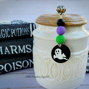 May include: A white ceramic jar with a wooden lid and a black and white ghost charm hanging from it. The charm is attached to a string with purple, green, and black beads. The jar is decorated with a white floral pattern. The jar is sitting on a white surface with a stack of black books that say "Magic Potions", "Charms", and "Poison" in white lettering.