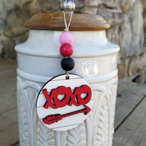 May include: A white ceramic jar with a wooden lid and a wooden round ornament with the word "XOXO" in red with a red heart and an arrow. The ornament is hanging from the lid with a string and pink, red, and black beads.