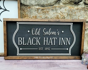 Puede incluir: Un letrero de madera negro y blanco con el texto "Old Salem's Black Hat Inn Est. 1892" en una fuente blanca de estilo manuscrito. El letrero está enmarcado en madera oscura y tiene un borde blanco.