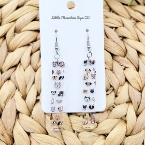 May include: A pair of silver dangle earrings with clear rectangular charms featuring a pattern of various dog breeds. The charms are printed with a variety of dog breeds in different colors. The earrings are labeled "Little Mountain Sign CO".