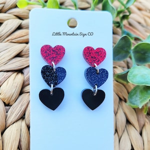 May include: A pair of dangle earrings with three layered hearts. The top heart is red glitter, the middle heart is dark blue glitter, and the bottom heart is black.