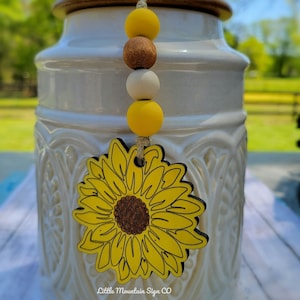 May include: A white ceramic jar with a brown lid and a sunflower-shaped wooden ornament hanging from it. The ornament is yellow with a brown center and has the text "Little Mountain Sign CO" at the bottom.
