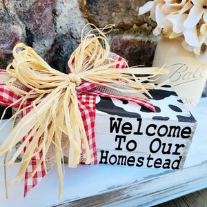 Puede incluir: Un letrero de madera con un diseño de vaca en blanco y negro. El letrero dice "Bienvenido a nuestra granja" y está decorado con una cinta de cuadros rojos y blancos y un lazo de paja.